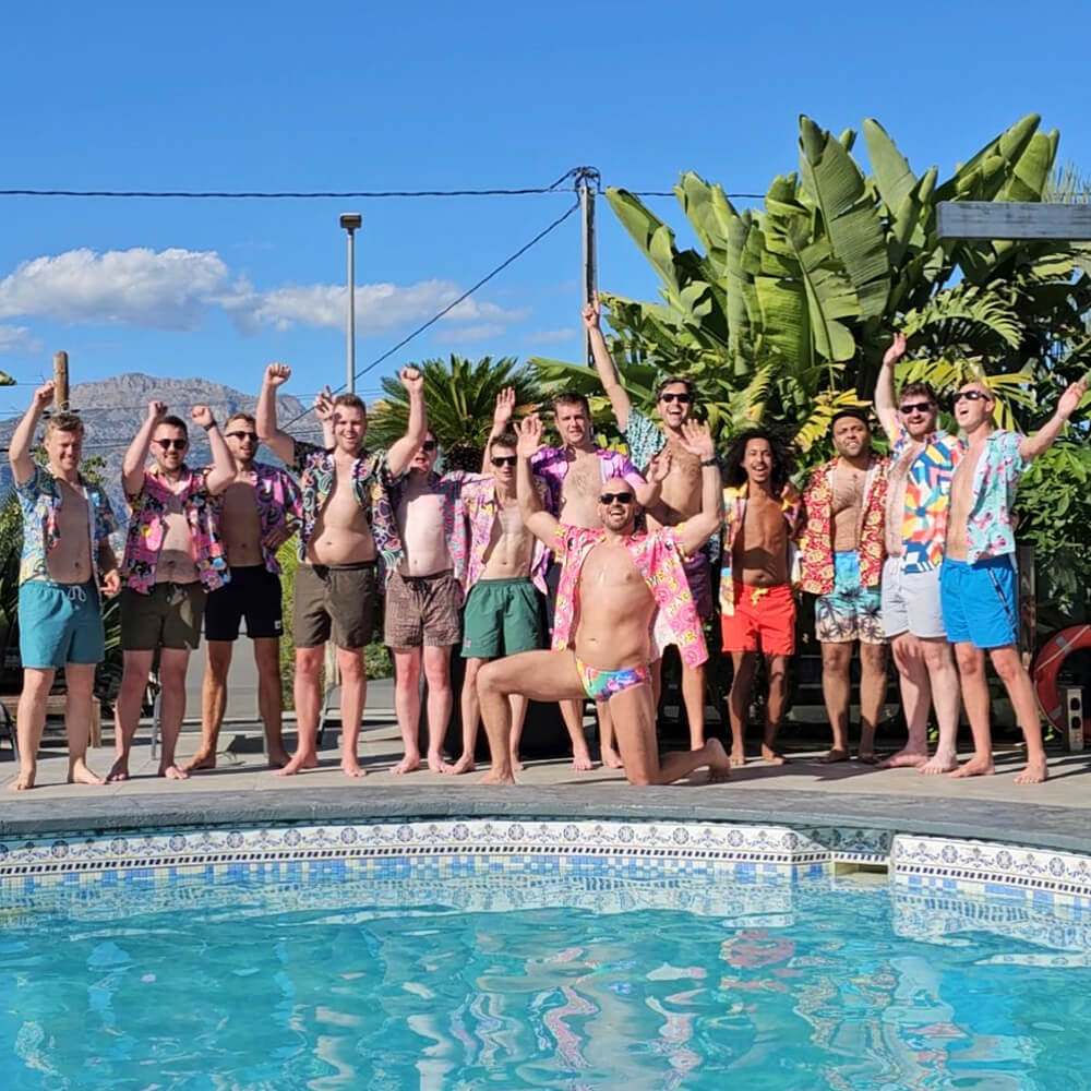 A group of men on a stag do in colourful shit shirt and swimwear stand and pose by a swimming pool on a sunny day, raising their arms and smiling, with lush green plants and a clear blue sky in the background.