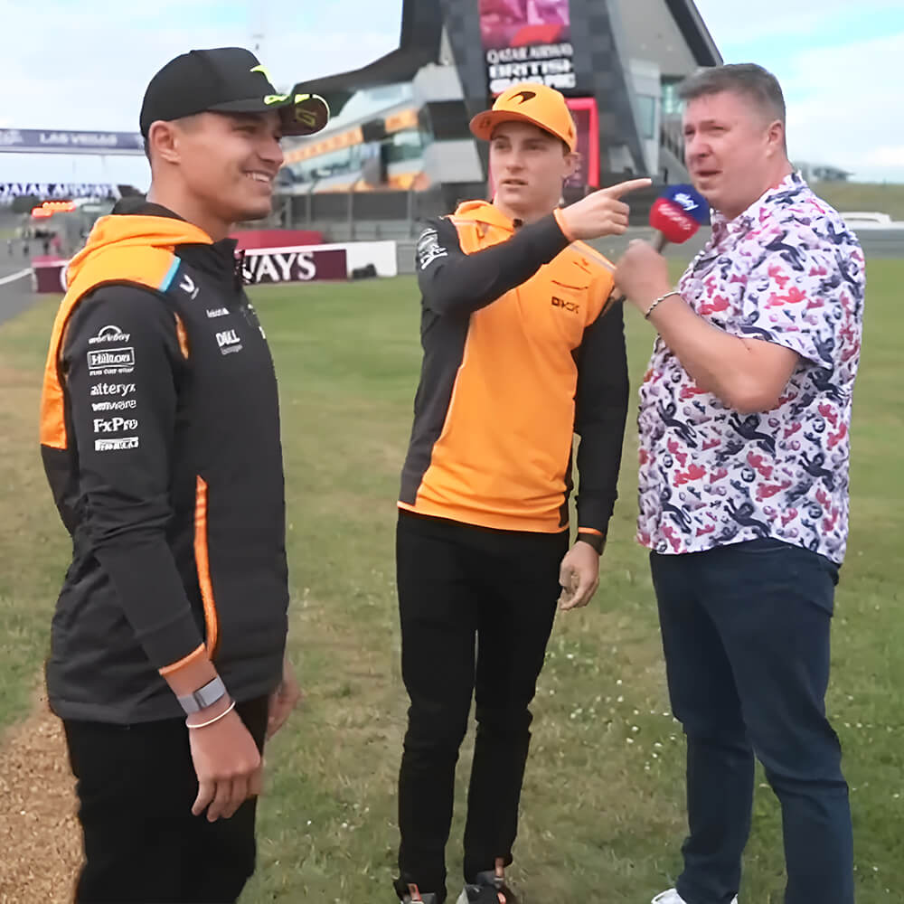 F1 commentator David Croft, also known as Crofty, wearing the England Shit Shirt holding a Sky Sports F1 microphone interviewing the McLaren drivers, Lando Norris and Oscar Piastri before the penalty shootout challenge.