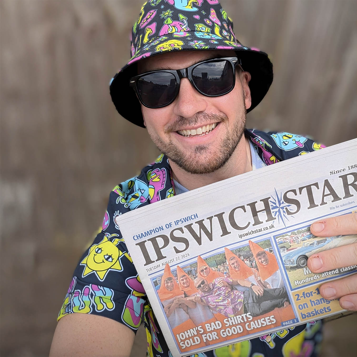 Shit Shirt Club founder in sunglasses and a colourful, shit shirt and matching bucket hat holds up a copy of the Ipswich Star newspaper featuring a front-page story titled 'John's Bad Shirts Sold for Good Causes'.