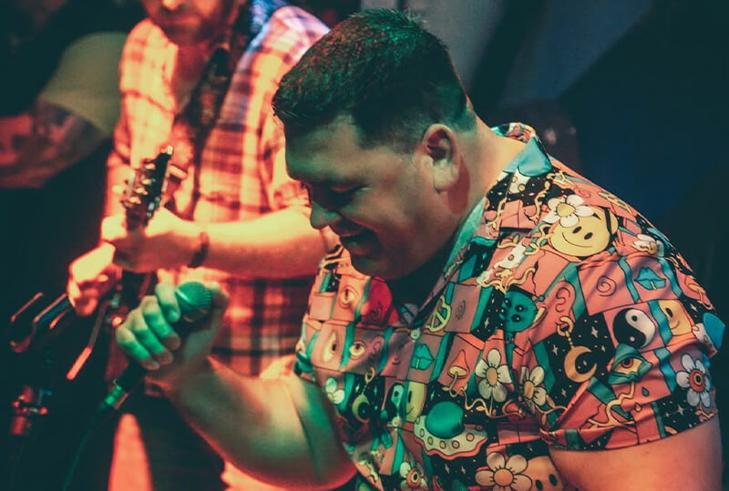 Man singing on stage wearing a colourful psychedelic print shit shirt from Shit Shirt Club during a live music performance.