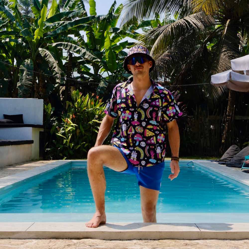 A man in the vibrant Shroom Shirt, paired with blue shorts and a bucket hat, strikes a confident pose with one foot on the pool edge amid lush tropical greenery and palm trees.