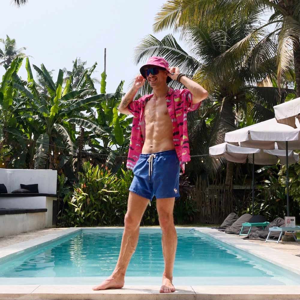 A man stands by a pool wearing the Movember Shirt in pink pattern, blue swim shorts, and a matching bucket hat. Palm trees and greenery surround the pool, with lounge chairs visible in the background.