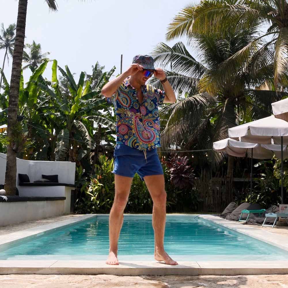 A man in a colorful shirt, blue shorts, and the Psychedelic Explorer Bucket Hat stands at a swimming pool’s edge, surrounded by tropical plants and palm trees, with lounge chairs and umbrellas visible in the background.