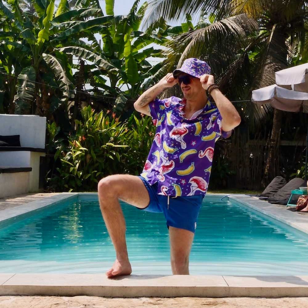 A man wearing a vibrant Bananas For Flamingos Shirt poses with one knee up by a sunny pool, surrounded by lush tropical plants and palm trees.