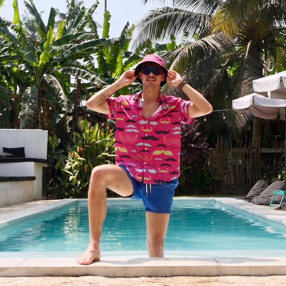 A man wearing a pink Movember Shirt with mustache patterns, blue shorts, and a bucket hat poses by a pool amid tropical plants and palm trees—showcasing the Movember Shirt as a stylish statement piece.