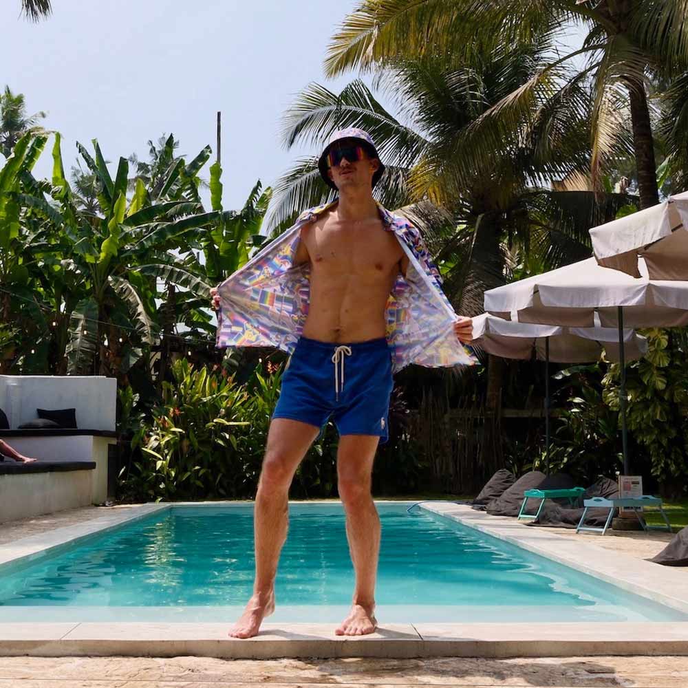 A man wearing an LGBTQ Pride Shirt, blue swim shorts, sunglasses, and a bucket hat stands confidently by a pool with tropical plants and palm trees in the background.