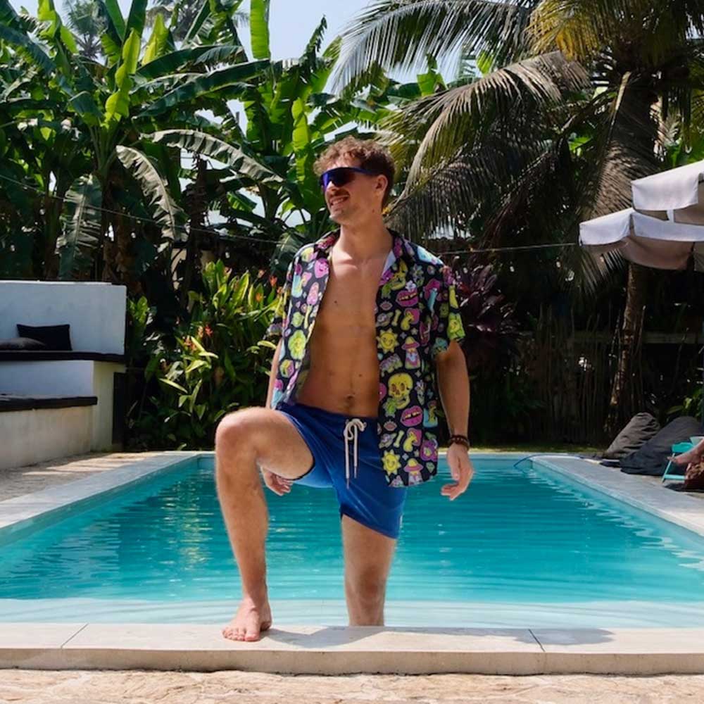 A man in the Black Out Mushroom Shirt, sunglasses, and blue swim trunks poses with one knee on the pools edge, surrounded by palm trees and greenery under bright sunlight for a vibrant festival-ready look.