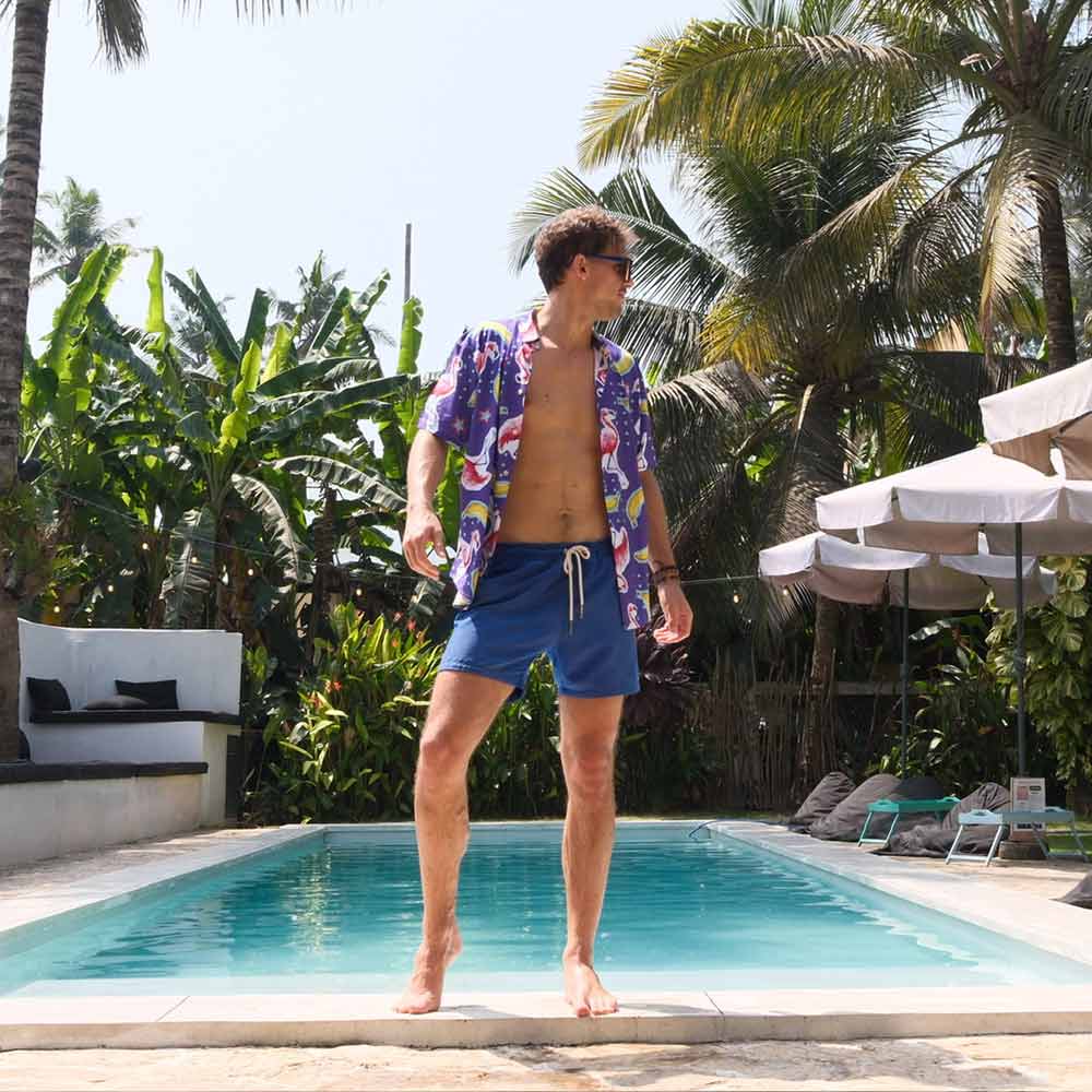 A man in the Bananas For Flamingos Shirt and blue swim shorts stands poolside amid tropical plants, palm trees, lounge chairs, and umbrellas.