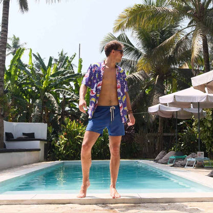 A man in the Bananas For Flamingos Shirt and blue swim shorts stands poolside amid tropical plants, palm trees, lounge chairs, and umbrellas.