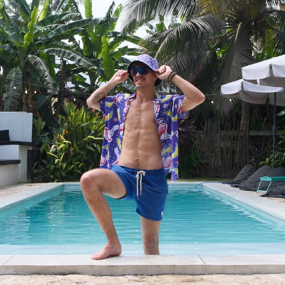 A man in sunglasses, blue shorts, and a matching hat poses by a pool with one knee up, surrounded by tropical plants and palm trees while wearing the Bananas For Flamingos Shirt.