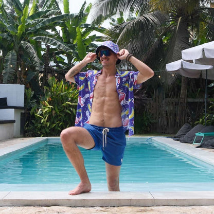 A man in sunglasses, blue shorts, and a matching hat poses by a pool with one knee up, surrounded by tropical plants and palm trees while wearing the Bananas For Flamingos Shirt.