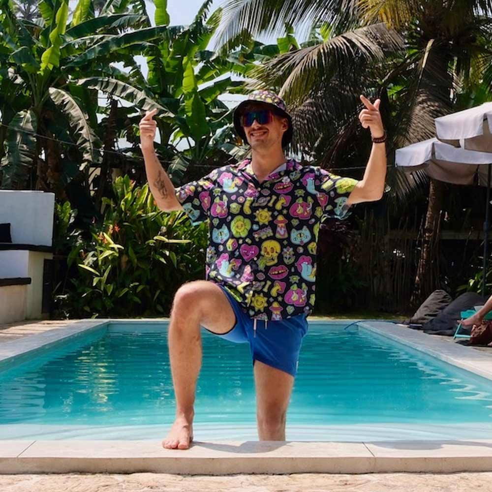 Wearing the Black Out Mushroom Shirt, blue shorts, and a bucket hat, a person poses poolside with one knee up, smiling and pointing skyward—surrounded by tropical plants and palm trees—for the perfect festival fashion vibe.