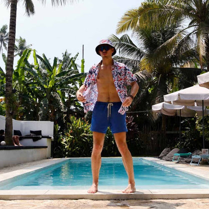 A man in blue shorts, sunglasses, a cap, and the England Football Hawaiian Shirt stands confidently by a swimming pool lined with palm trees and lounge chairs on a sunny day.