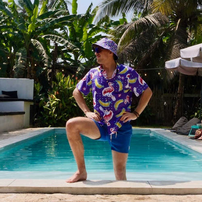Wearing the Bananas For Flamingos Bucket Hat, a person in a colorful shirt and blue shorts poses with one foot on the pool edge, surrounded by tropical plants and palm trees in a sunny outdoor pool setting.
