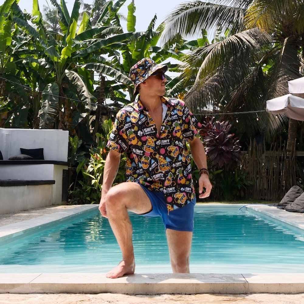 Wearing the Wanna Pizza This Shirt, a man pairs it with a matching pizza bucket hat and blue shorts, posing confidently poolside amid tropical greenery and palm trees.