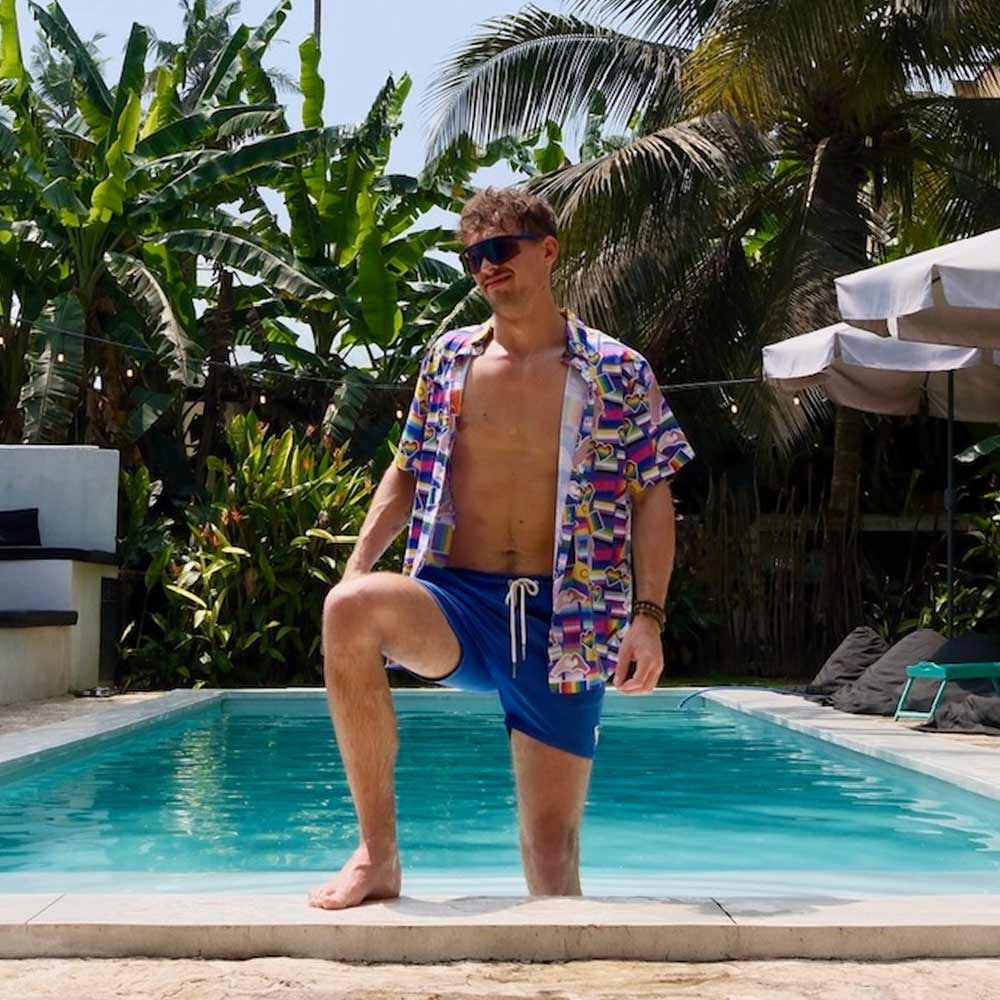 Wearing sunglasses and an LGBTQ Pride Shirt, a man in blue swim trunks stands poolside with one foot in the water, surrounded by tropical plants and palm trees, looking to the side.