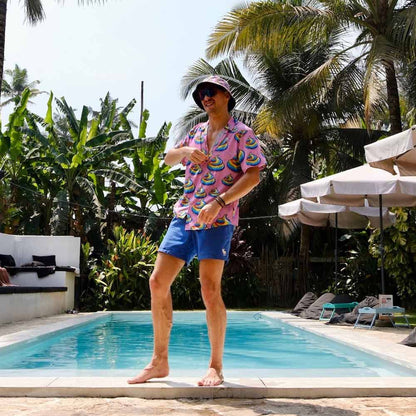 Wearing a colorful shirt, blue shorts, and the Unicorn Poo Bucket Hat, a person stands playfully by a pool, surrounded by lush tropical plants and palm trees on a sunny day.