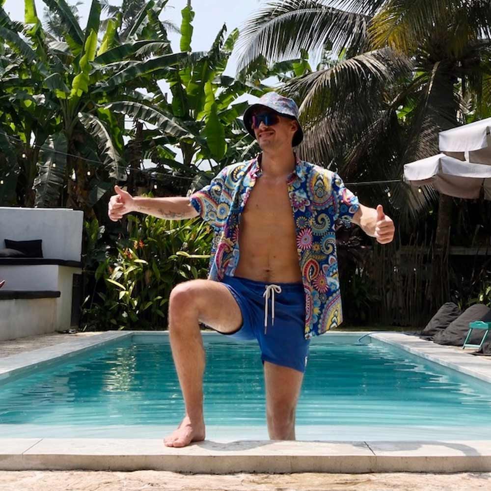 A man in a colorful shirt, blue shorts, and the Psychedelic Explorer Bucket Hat stands at the edge of a pool, giving two thumbs up as tropical plants and palm trees surround the area.