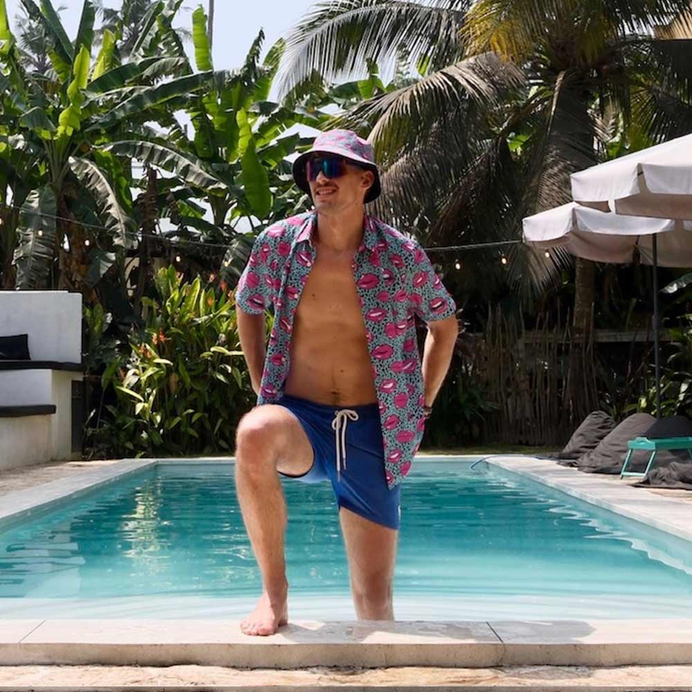 Wearing the Leopard Kiss Bad Shirt with playful feline patterns, a man in a colorful bucket hat and sunglasses stands poolside, one knee raised, amid lush tropical plants and palm trees on a sunny day.