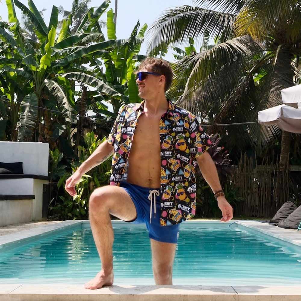 A man wearing sunglasses, a “Wanna Pizza This Shirt,” and blue swim shorts stands in a pool with one knee raised, surrounded by palm trees and lush greenery.