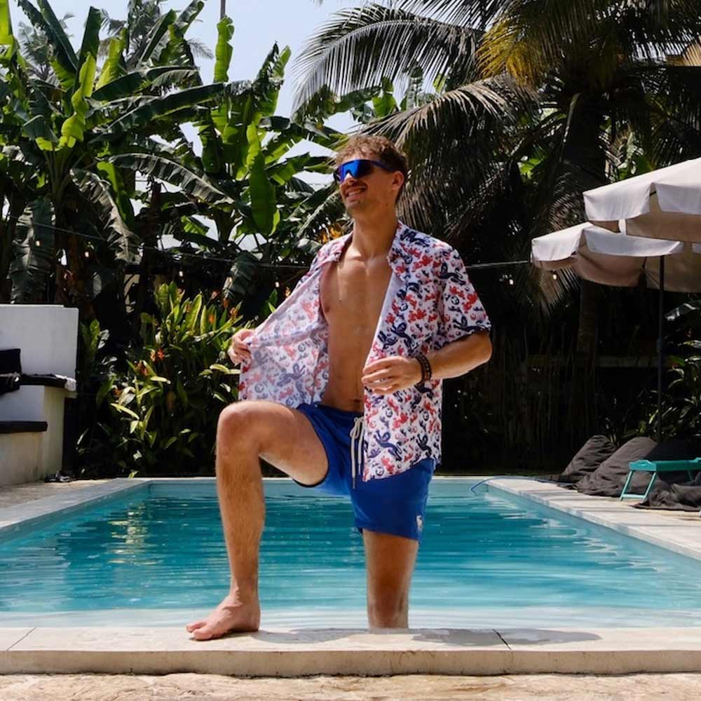 A man in sunglasses and blue swim trunks, wearing an unbuttoned England Football Hawaiian Shirt, stands with one knee on a pool edge surrounded by lush tropical plants and palm trees.