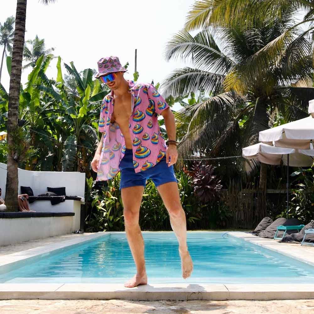 A man in a vibrant Unicorn Poo Bucket Hat, an open rainbow-print shirt, and blue shorts walks by a pool surrounded by palm trees and lounge chairs on a sunny day.