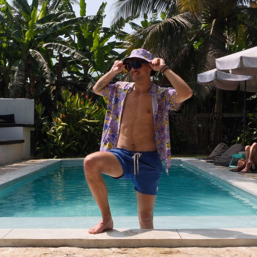 Wearing the Magical Psychedelic Shirt, a man in blue swim trunks and a matching bucket hat poses confidently by a pool, surrounded by lush tropical plants and palm trees on a sunny day.