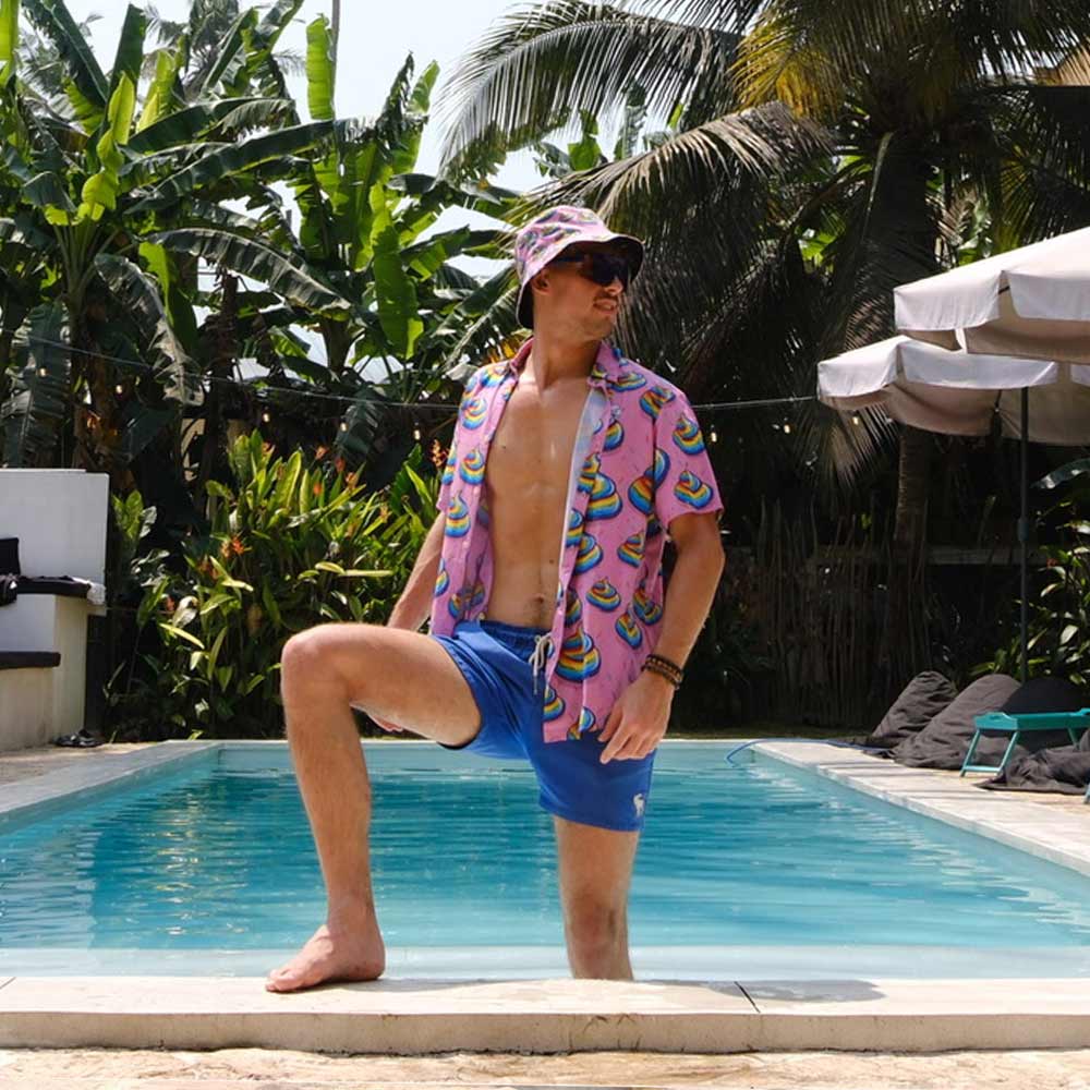 Wearing the Unicorn Poo Bucket Hat, a man in blue swim trunks and a patterned pink shirt poses by the poolside with one foot on the edge, surrounded by lush tropical plants and palm trees.
