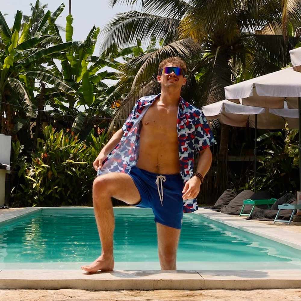A man in a Combat2Coffee Army Camo Shirt, sunglasses, and blue swim trunks stands poolside among tropical plants and palm trees, promoting mental health awareness for veterans.