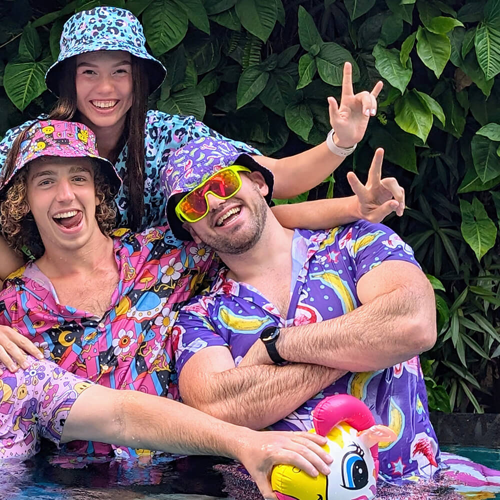 Three people in colorful, cartoon-themed outfits and bucket hats pose cheerfully in front of lush green leaves, smiling and making peace signs. One holds an inflatable unicorn pool toy.