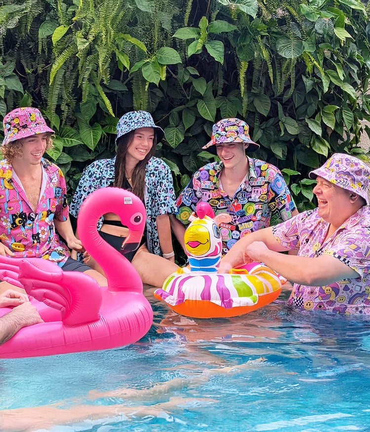 Four people wearing shit shirts and matching bucket hats in a pool with colourful inflatables and funny bucket hats, surrounded by greenery.