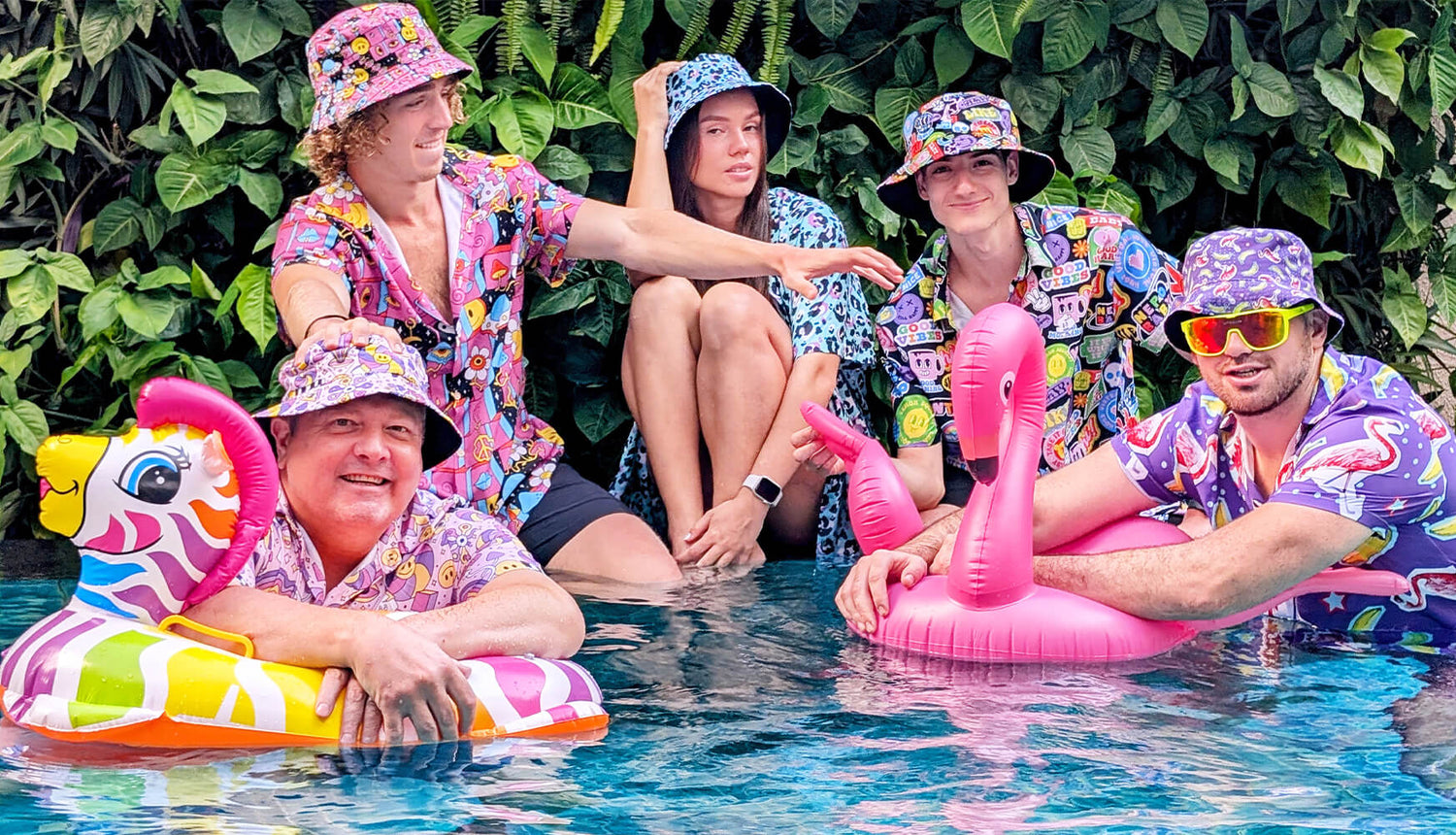 Five people in colorful shirts and bucket hats pose in a pool with inflatable toys, including a tiger and a flamingo, against a backdrop of green plants. Everyone appears relaxed and cheerful.