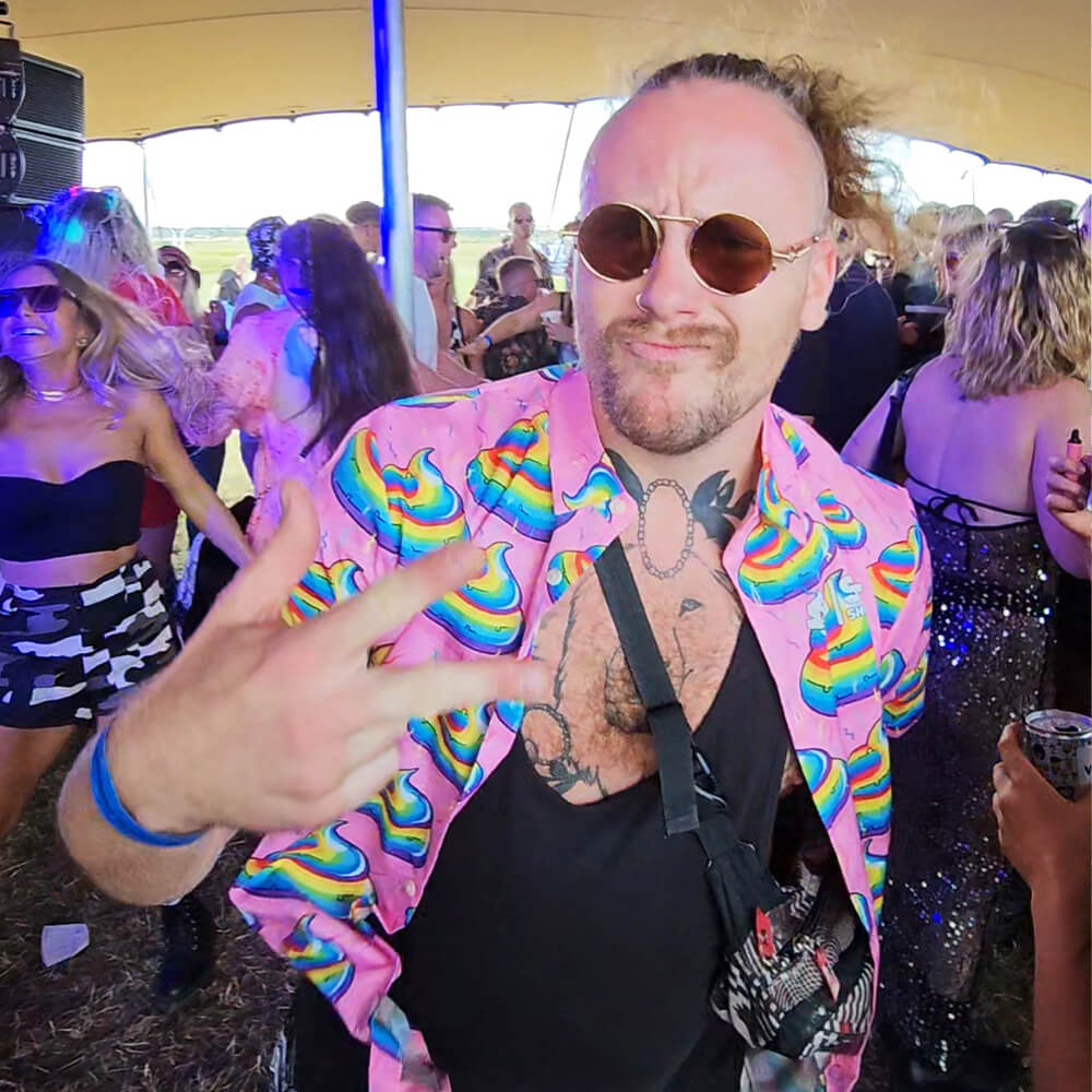 A man wearing round sunglasses and a colourful shit shirt with rainbow patterns makes a peace sign at an outdoor party, surrounded by dancing people under a canopy.