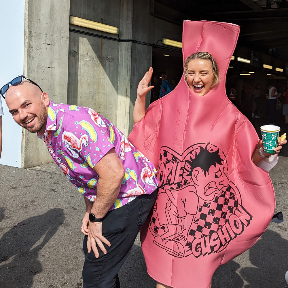 A man in a colourful flamingo shit shirt laughs as a woman dressed in a large whoopee cushion costume pretends to press on his backside; both are having fun Twickenham for the rugby sevens.