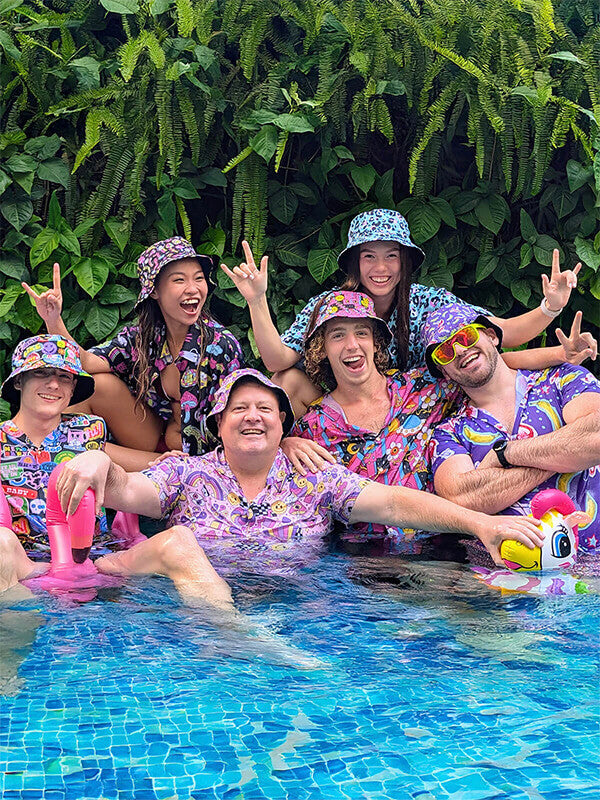 Group of people in a pool wearing funny bucket hats and bad shirts, surrounded by green foliage.