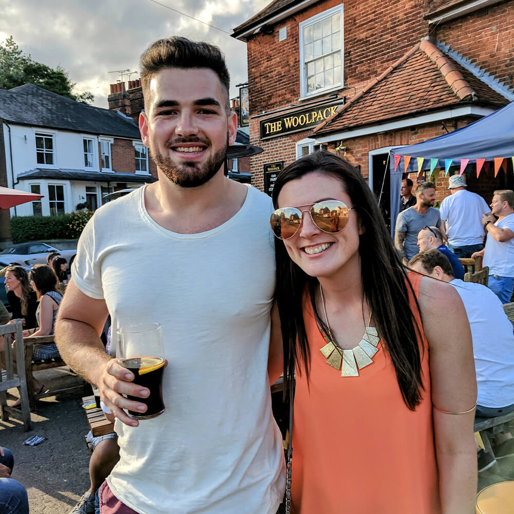 A smiling man with a beard holds a drink and stands next to a smiling woman in sunglasses and an orange top. They are outdoors at a lively pub garden with people and colourful bunting in the background.