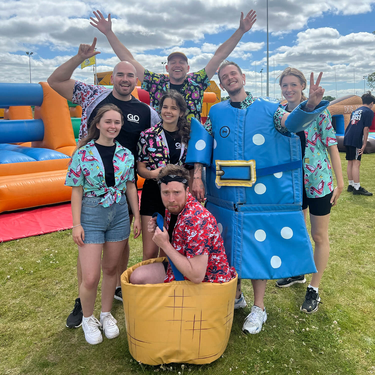 A group of seven people pose outdoors on grass, some wearing colourful shirts with fun patterns. One person is in a blue costume shaped like a vest, and another crouches in a basket costume. Inflatable obstacle courses are in the background.