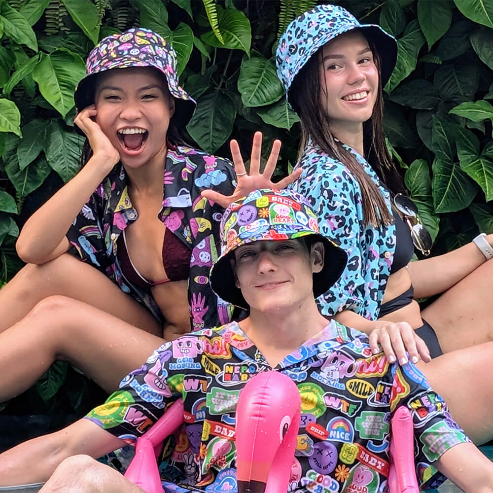 Three friends in bold Bad Shirt Subscription tops and bright bucket hats pose joyfully by leafy greenery. One woman smiles with her hand raised, while the others relax on a pink flamingo float, celebrating friendship and mental health awareness.