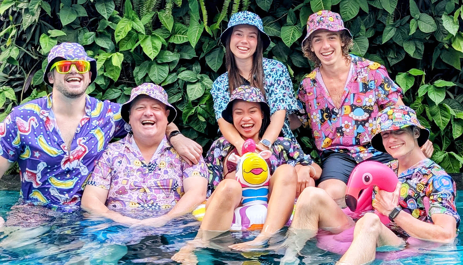 Six people wearing colourful shit shirts and bucket hats smile and pose together in a pool surrounded by green plants, with two inflatable toys—a duck and a flamingo—in front of them.