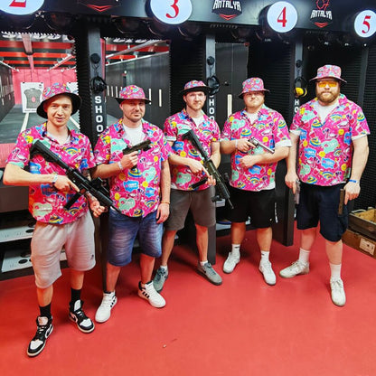 Five men at a shooting range pose with rifles, all wearing matching Badass Bucket Hats and colorful shirts that coordinate with the range’s red and black color scheme.