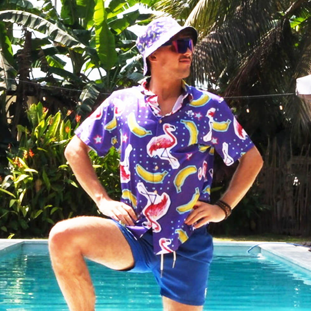 Wearing a Bananas For Flamingos Shirt, matching bucket hat, and blue shorts, a person confidently poses by a pool amid lush tropical plants.