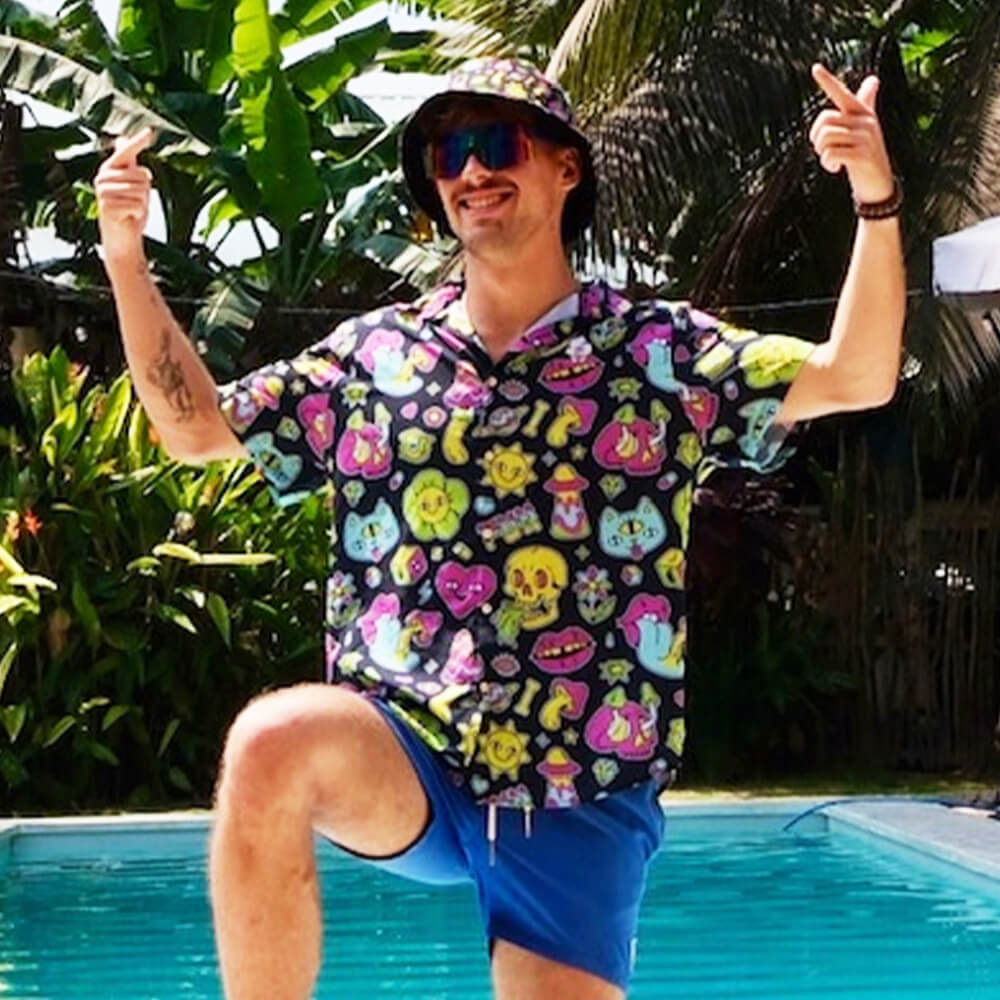 A smiling man in the Black Out Mushroom Shirt, blue shorts, and a bucket hat gives two thumbs up by a pool with lush tropical plants and palms—a fun festival fashion look.