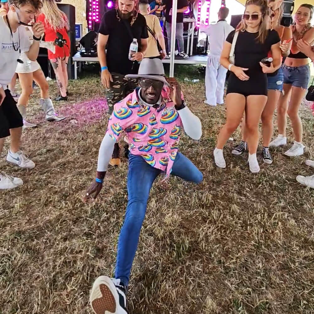 A man in a colourful shit shirt and hat is performing an energetic dance move on grass at an outdoor event, surrounded by people cheering and smiling.