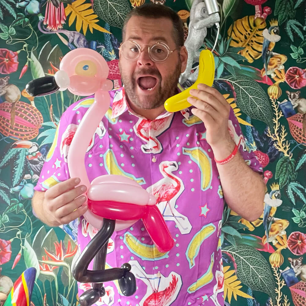 A man in a pink flamingo-patterned shit shirt holds a balloon flamingo and a yellow balloon shaped like a banana, standing in front of a colourful, tropical-patterned background.