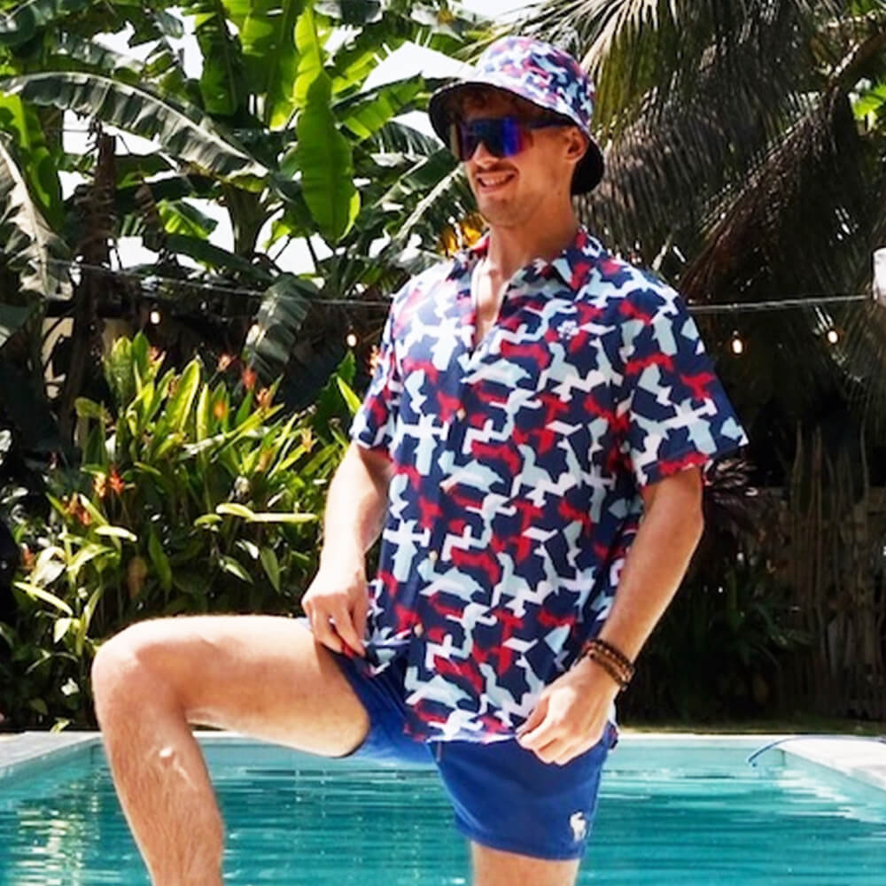 Wearing a Combat2Coffee Army Camo Shirt, a person stands by the pool amid palm trees, smiling in the sun. Their vibrant style supports Combat2Coffee and raises awareness for veterans mental health.