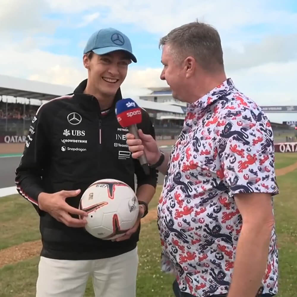 George Russell in a Mercedes jacket and blue cap holds a soccer ball while being interviewed by Crofty in a colourful England shit shirt holding a Sky Sports microphone at Silverstone Circuit.