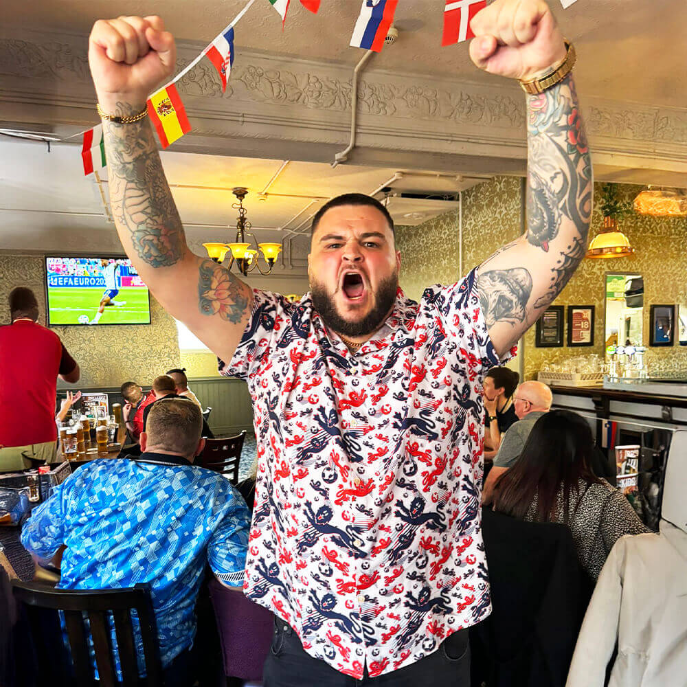 Man wearing the England Football Shit Shirt with tattoos celebrating in a bar with the Euros on TV