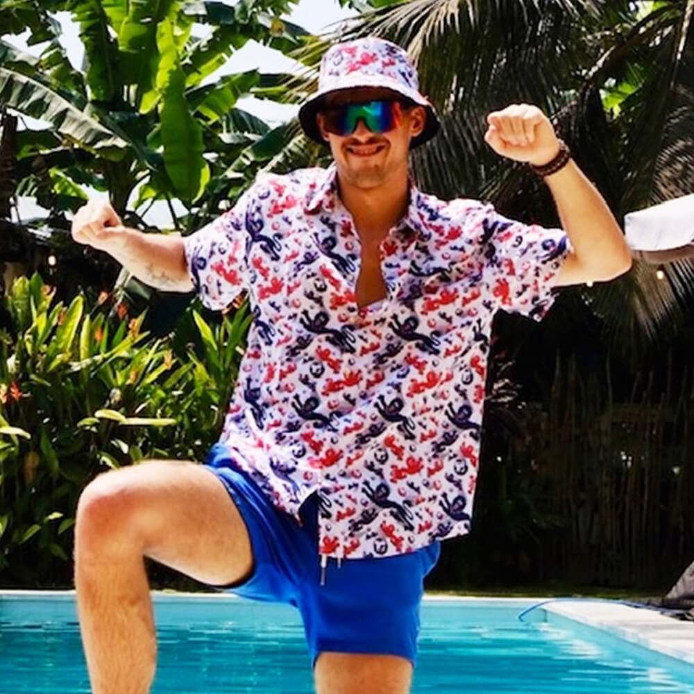 A man in sunglasses, blue shorts, and the England Football Hawaiian Shirt strikes a playful pose by a pool surrounded by lush tropical plants, showing off his fun summer style.
