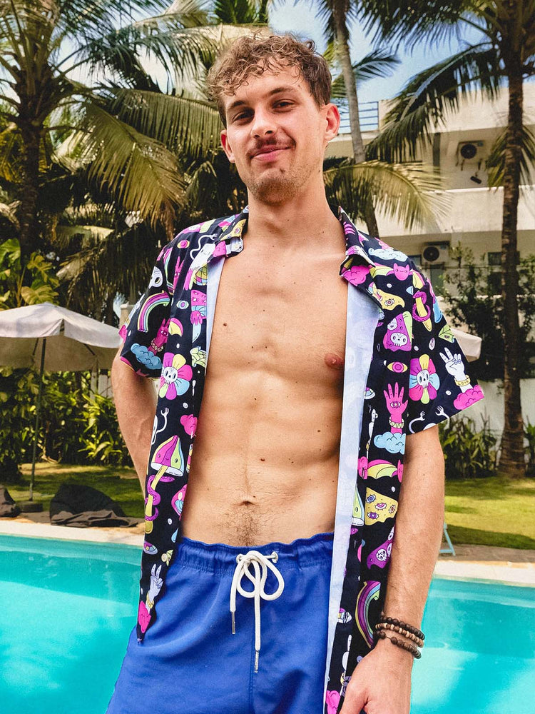 A young man with curly hair stands by a pool, wearing blue swim shorts and an open, colourful short-sleeve shit shirt with cartoon designs. Palm trees and a modern building are in the background.