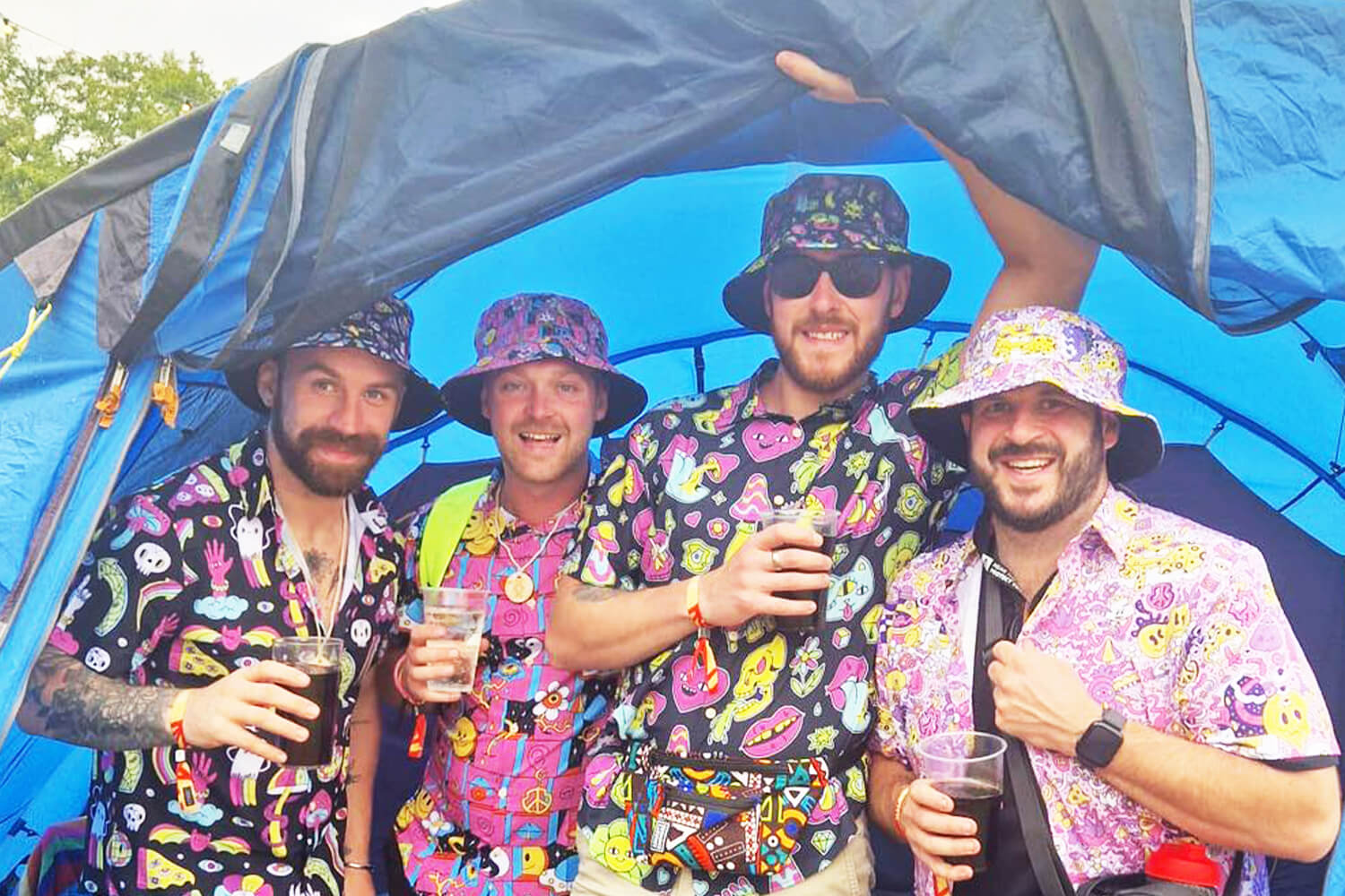 Four smiling men stand together in front of a blue tent, wearing colourful patterned shit shirts and matching bucket hats. They are each holding drinks and appear to be enjoying a music festival.